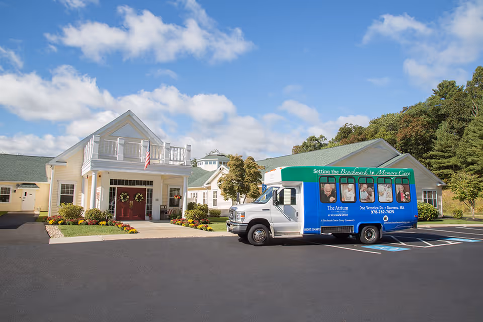 Front entrance of a yellow senior living facility with landscaping and a blue branded shuttle van parked in the driveway under a partly cloudy sky.