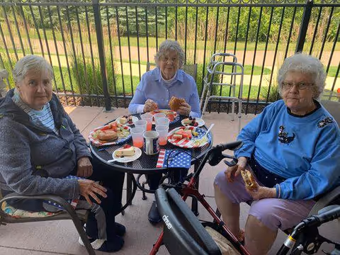 Three elderly women sitting around a round outdoor table on a patio, enjoying a meal with plates of sandwiches, watermelon, and drinks. Two of the women have walkers nearby. The background shows a fenced grassy area with trees.