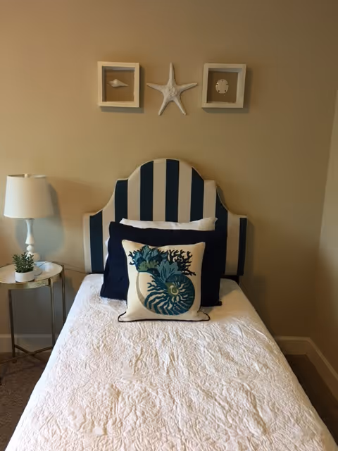 A neatly made single bed with a blue and white striped headboard, white quilted bedspread, and decorative pillows featuring sea-themed designs. Above the bed are three wall decorations: two framed seashells and a starfish in the center. To the left of the bed is a small round side table with a white lamp and a small potted plant.