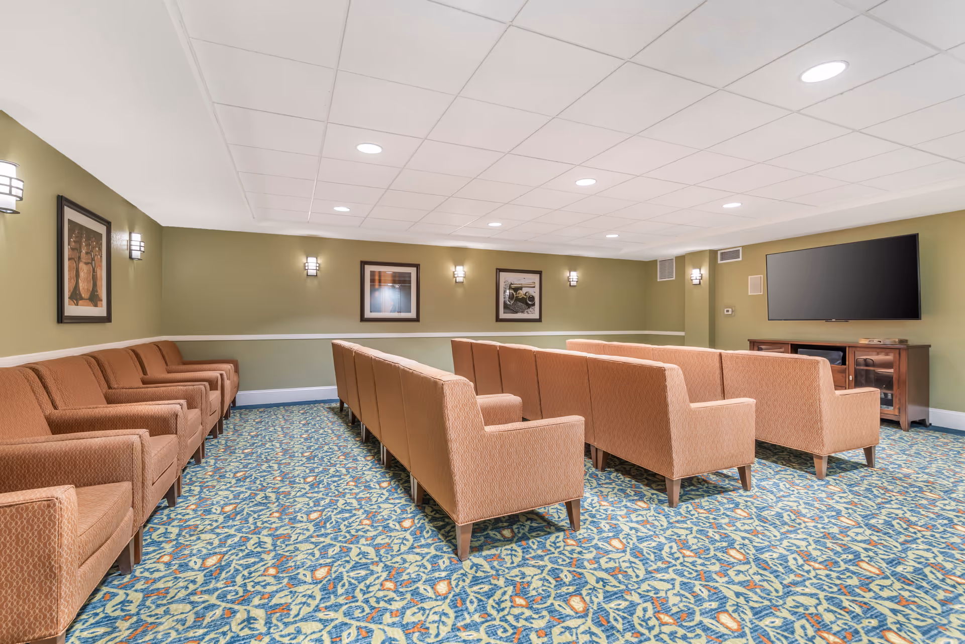 A spacious common room with rows of brown upholstered chairs facing a large flat-screen TV mounted on a green wall. The room has patterned carpet flooring, framed artwork on the walls, and ceiling lights providing bright illumination.