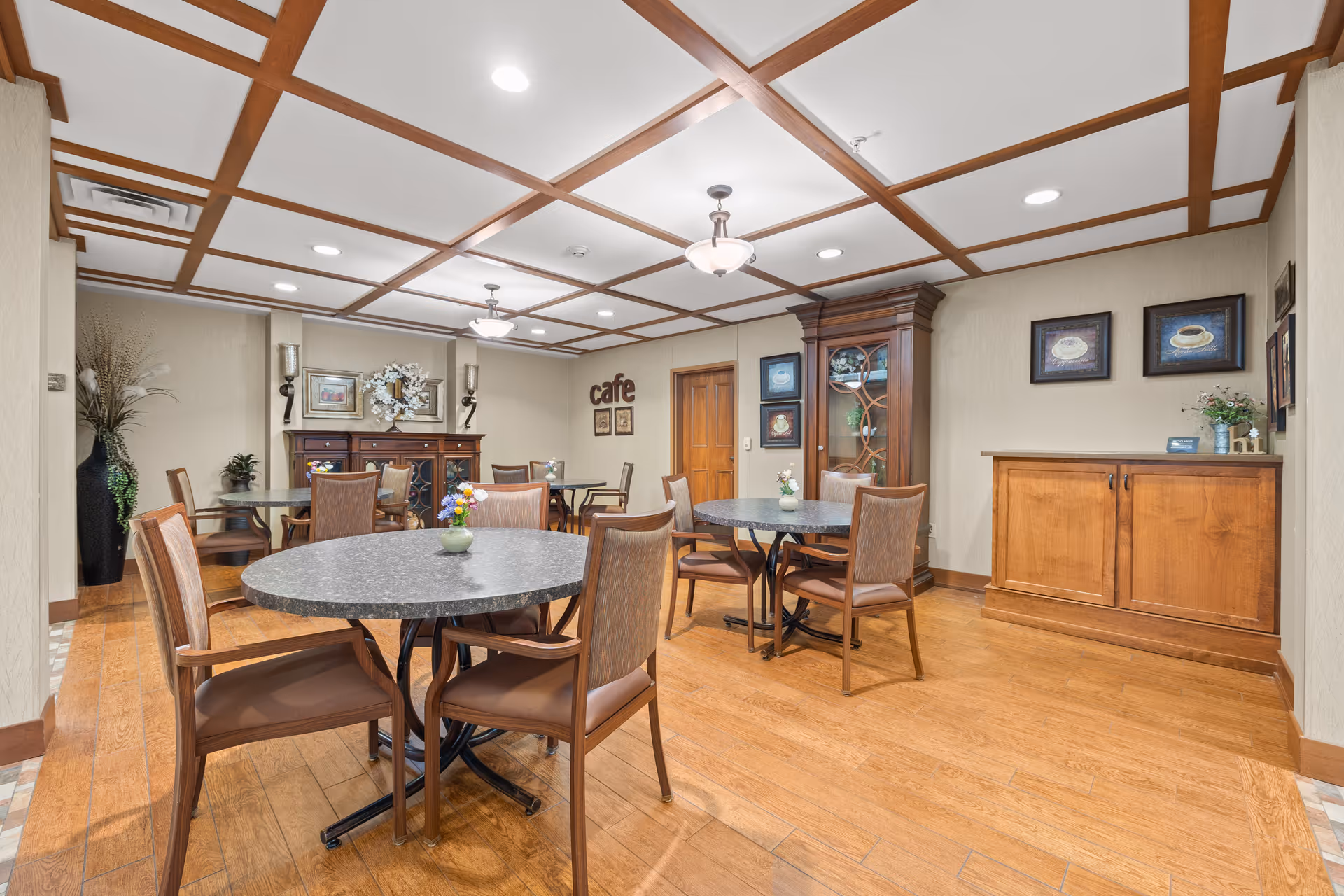A cozy dining area in a senior living facility with round tables and wooden chairs arranged neatly. The room features a wooden coffered ceiling, hardwood floors, and warm beige walls. Decorative elements include framed pictures, a wooden cabinet, a sideboard, and a large vase with plants. The word 'cafe' is displayed on one wall.