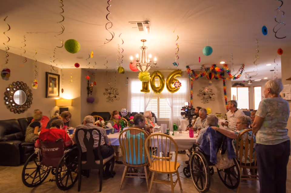A group of elderly people gathered around a dining table in a decorated room celebrating a 100th birthday with gold balloons shaped as '100'. The room is festively adorned with hanging streamers and colorful decorations. Some attendees are seated in wheelchairs, and others are sitting on wooden chairs. A chandelier hangs from the ceiling, and there is a cameraman recording the event.