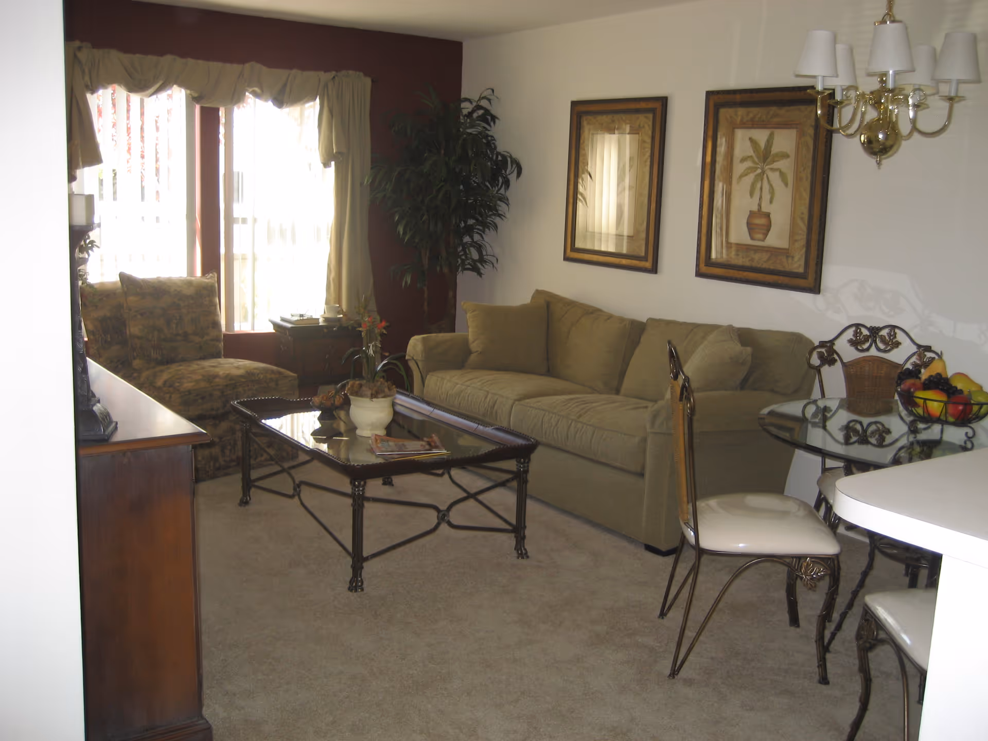 A cozy living room area with a beige sofa, a glass coffee table with decorative items, a patterned armchair near a window with curtains, two framed botanical prints on the wall, a glass dining table with metal chairs, and a chandelier overhead.