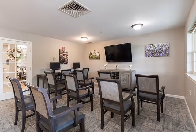 A small meeting or activity room with several wooden chairs arranged in rows facing a wall-mounted flat screen TV. Below the TV is a wooden cabinet with decorative flowers on top. The room has light-colored walls with three framed floral pictures and a window letting in natural light. There is a door with glass panels leading to another room with a dining table and chairs.