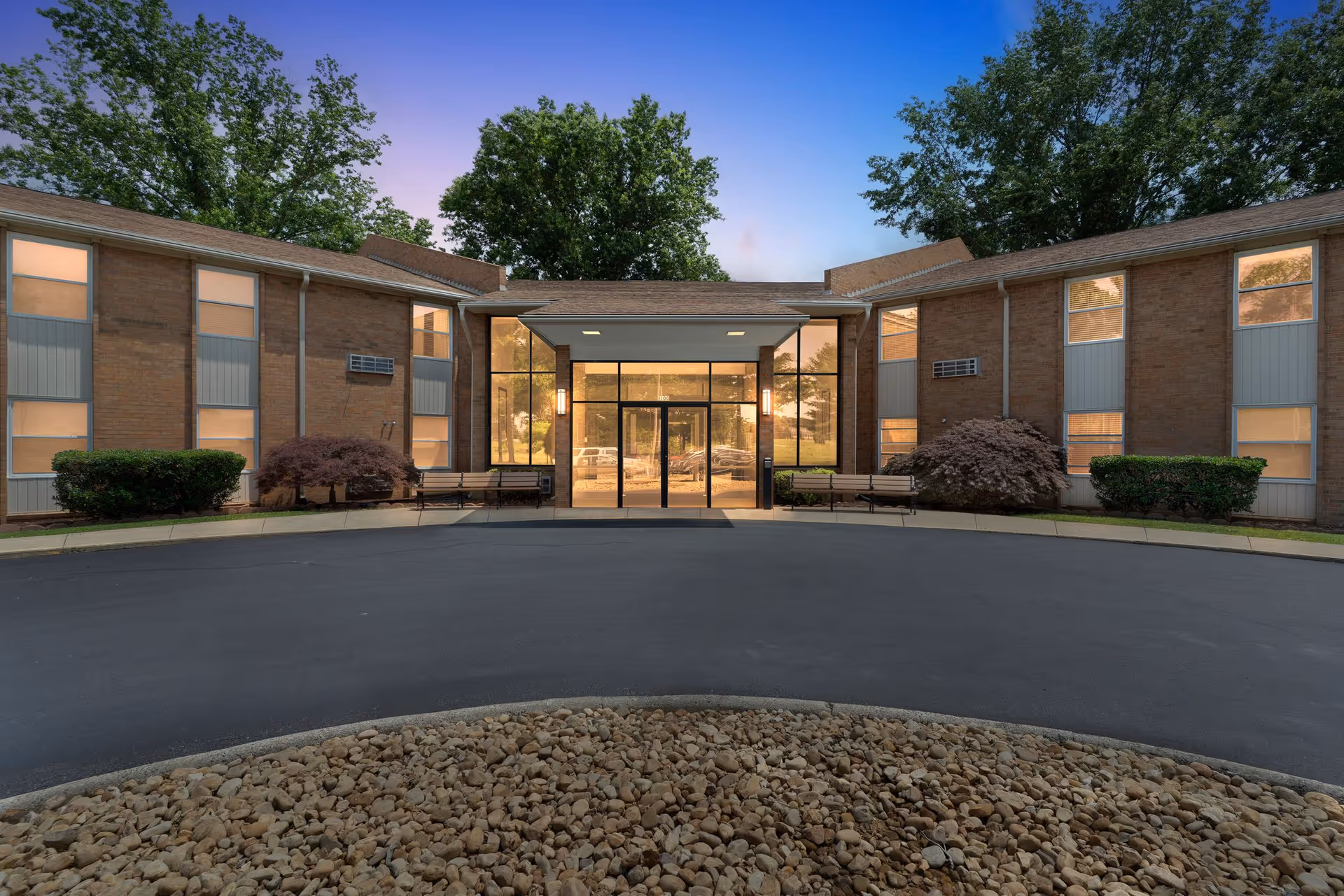Front exterior of a brick senior living building featuring a glass entryway, benches, landscaping, and a circular driveway at dusk.