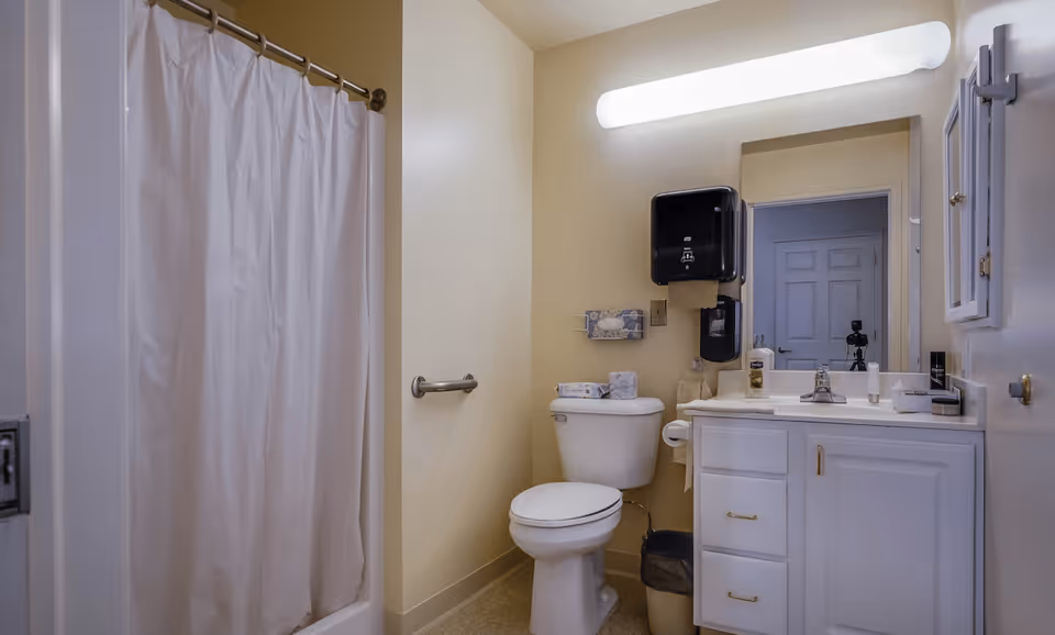 A clean bathroom with a white shower curtain, a toilet with a grab bar on the wall beside it, a white vanity with a sink, a large mirror above the sink, and a wall-mounted soap dispenser and paper towel holder. The walls are painted light beige and the floor has a light-colored tile.