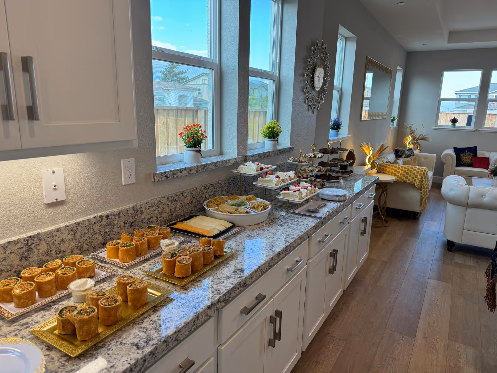 A bright and modern interior space with a long granite countertop displaying an assortment of appetizers and desserts. The countertop is lined with white cabinets and decorated with small potted plants on the windowsills. In the background, there is a cozy living area with white sofas, a yellow throw blanket, decorative pillows, a wall clock, and a large mirror. Large windows allow natural light to fill the room.
