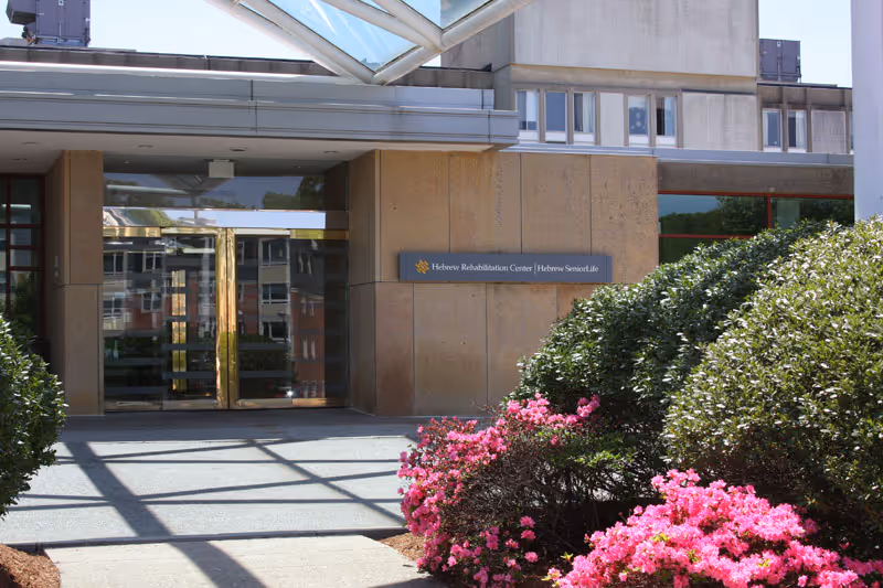 Entrance to the Hebrew Rehabilitation Center in Boston with glass doors, beige stone walls, and a sign displaying the facility's name. The entrance is surrounded by green bushes and pink flowering plants under a glass canopy.