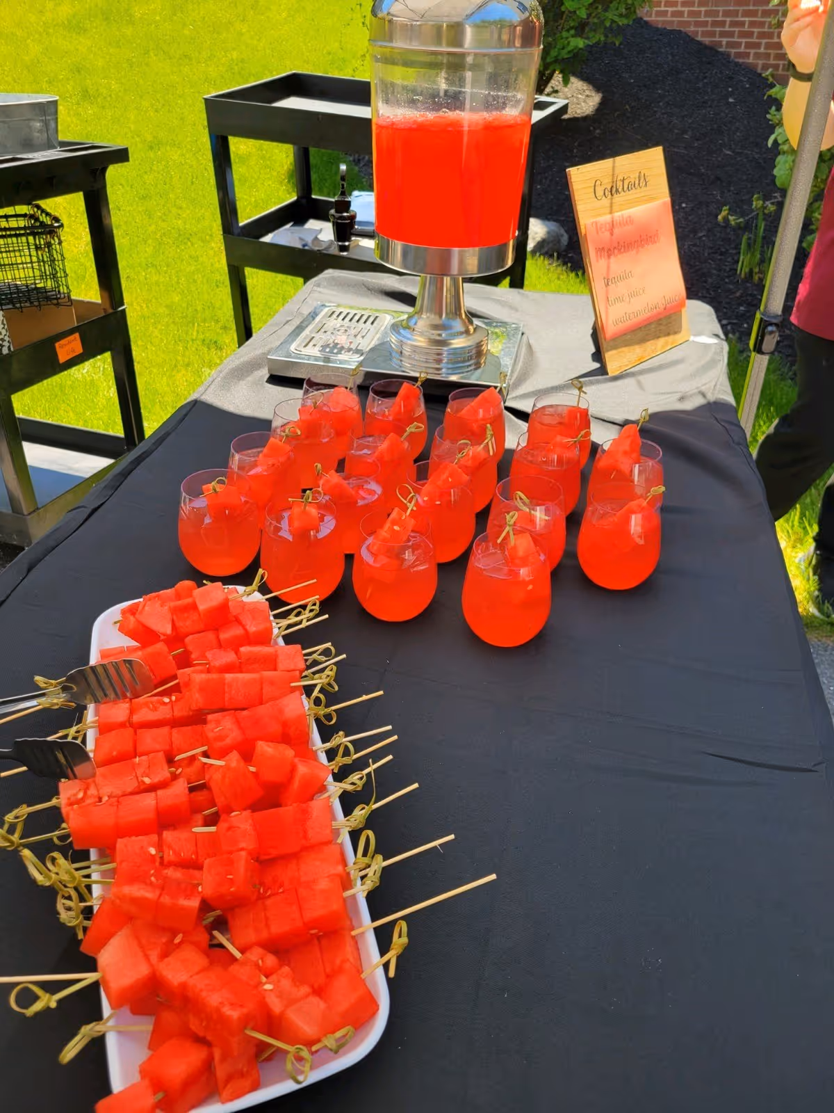 Outdoor table setup with a large beverage dispenser filled with red drink, multiple glasses of red cocktail garnished with watermelon cubes, and a tray of watermelon skewers. A small sign listing cocktail ingredients is also visible on the table.