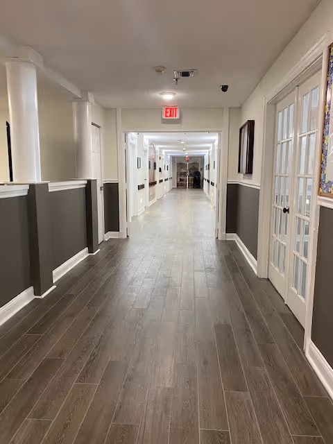 A long, well-lit hallway with wood flooring and white walls with gray wainscoting. There are several doors along the hallway, including a set of French doors on the right side. An exit sign is visible at the far end of the hallway.