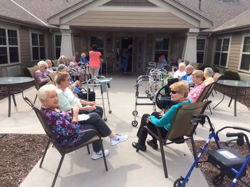 A group of elderly people sitting outside on chairs arranged in two rows facing each other on a concrete patio in front of a building entrance. Several walkers and wheelchairs are visible, and a staff member in a pink shirt is standing near the entrance. The setting appears to be a senior living facility.