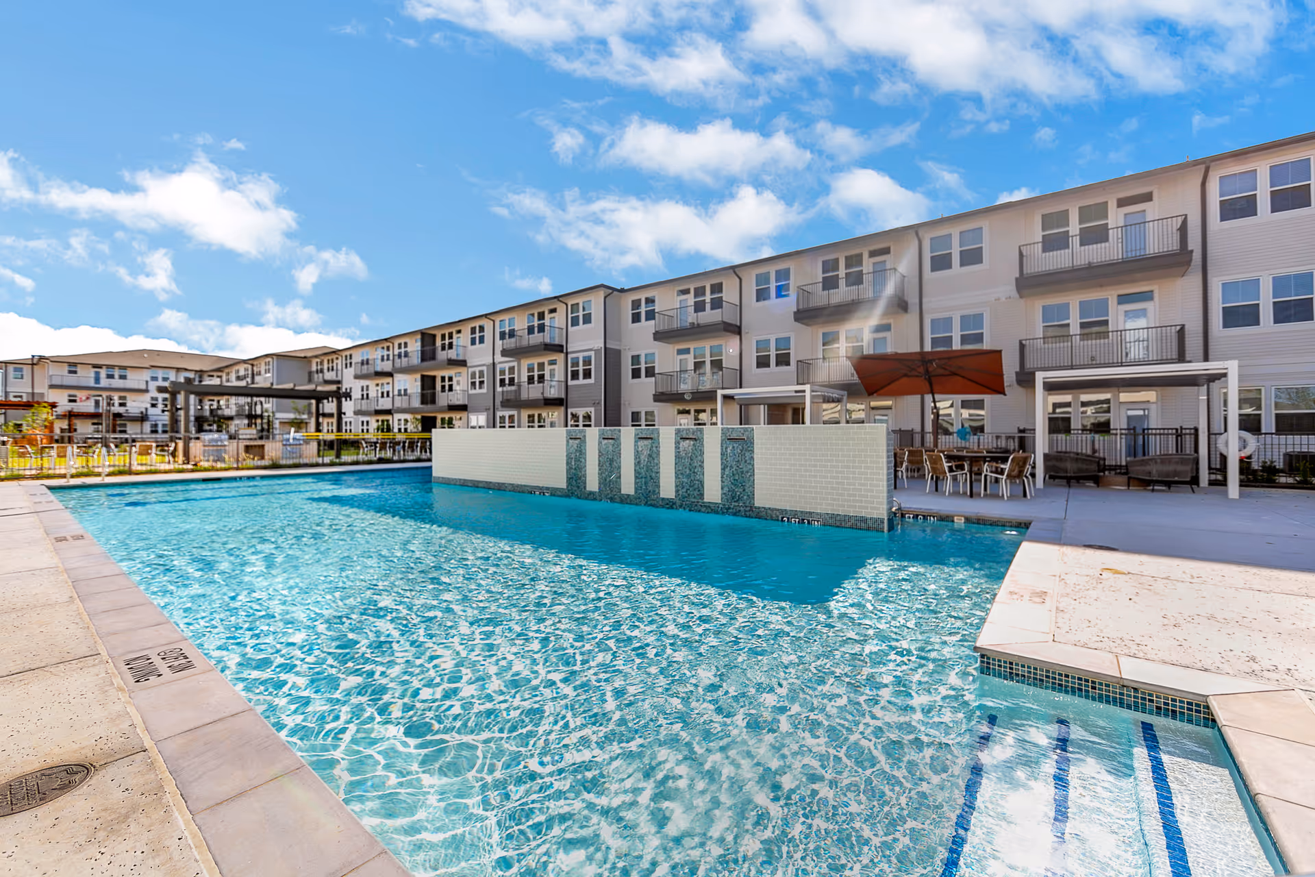 Outdoor swimming pool with clear blue water in front of a multi-story residential building under a partly cloudy sky. The pool area includes a tiled divider wall with water features, poolside seating with tables and chairs under umbrellas, and a concrete deck surrounding the pool.