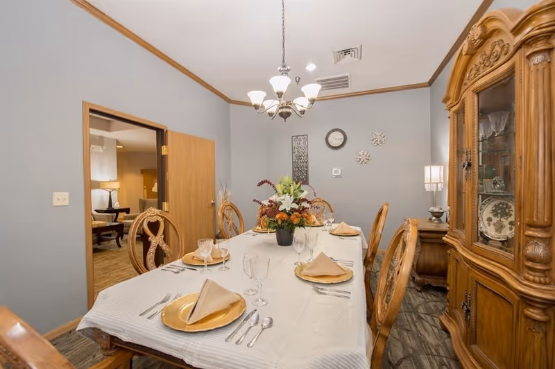 A formal dining room with an ornate wooden table set for six, a floral centerpiece, chandelier, and a china cabinet.