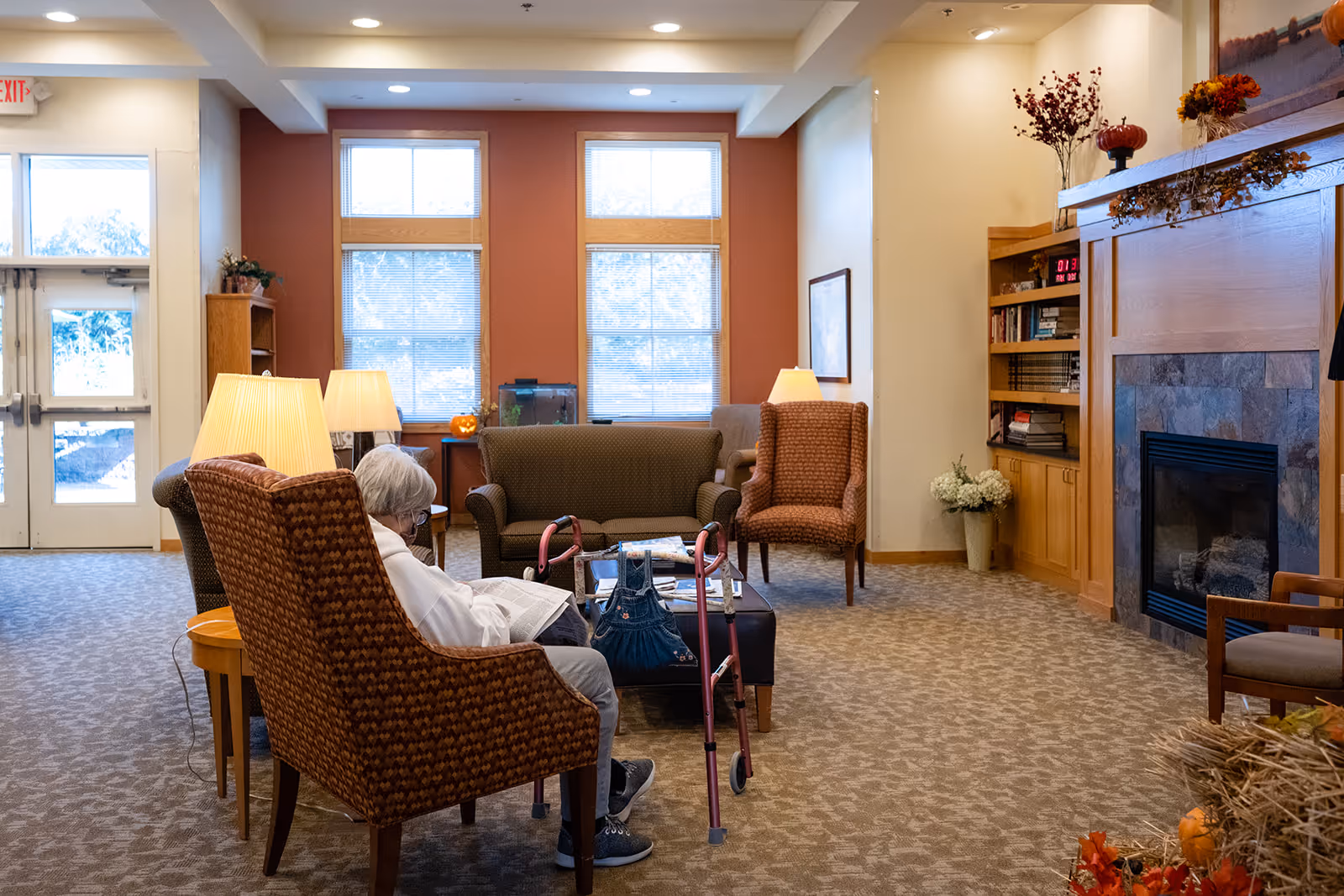 A person seated in a cozy lounge area of a senior living facility with armchairs, lamps, a fireplace, and a walker nearby.