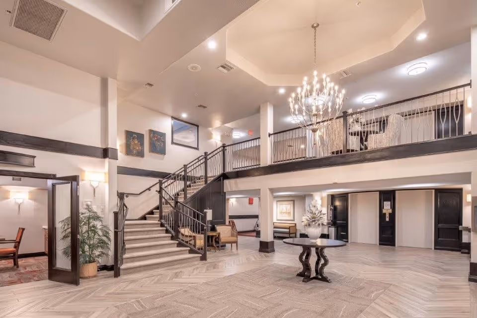Spacious and well-lit interior lobby area with a staircase leading to an upper level. The space features a large chandelier, a round table with a decorative vase, seating areas, and elevator doors. The decor includes light-colored walls, dark wood accents, and framed artwork.