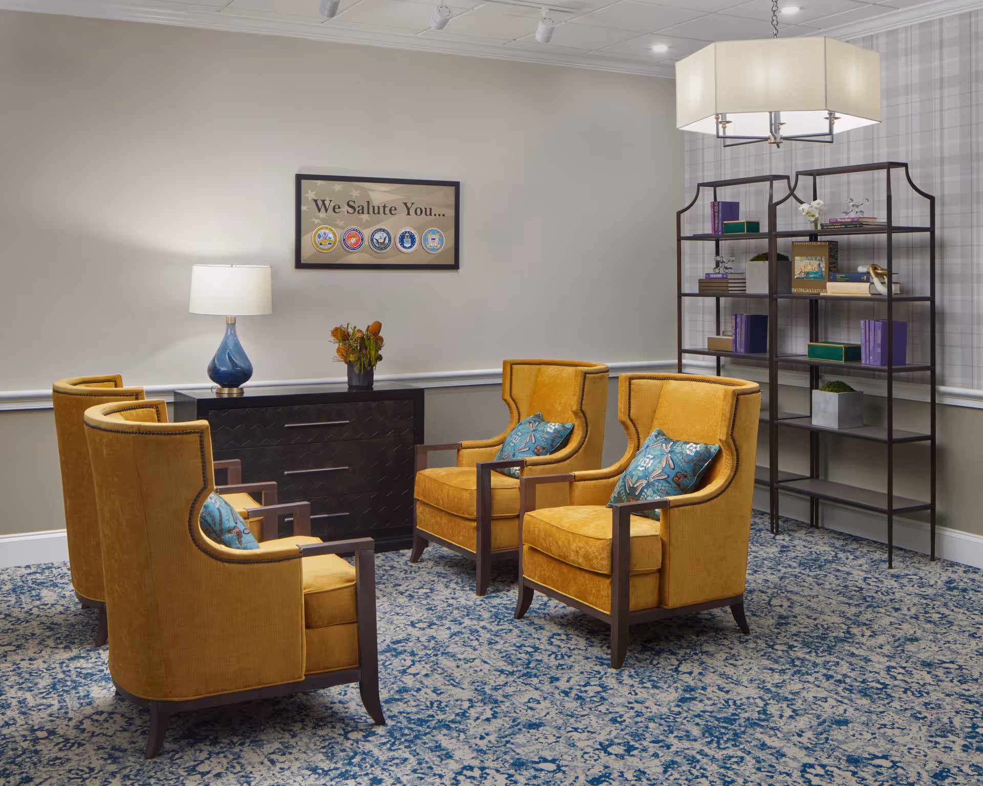 A cozy seating area with four mustard yellow armchairs arranged in a circle on a blue patterned carpet. A black cabinet with a blue lamp and a flower vase is against the wall. Above the cabinet, a framed sign reads 'We Salute You...' with military service emblems. To the right, a black metal bookshelf holds books and decorative items. The walls are light-colored with a plaid accent wall and a modern ceiling light fixture hangs above.