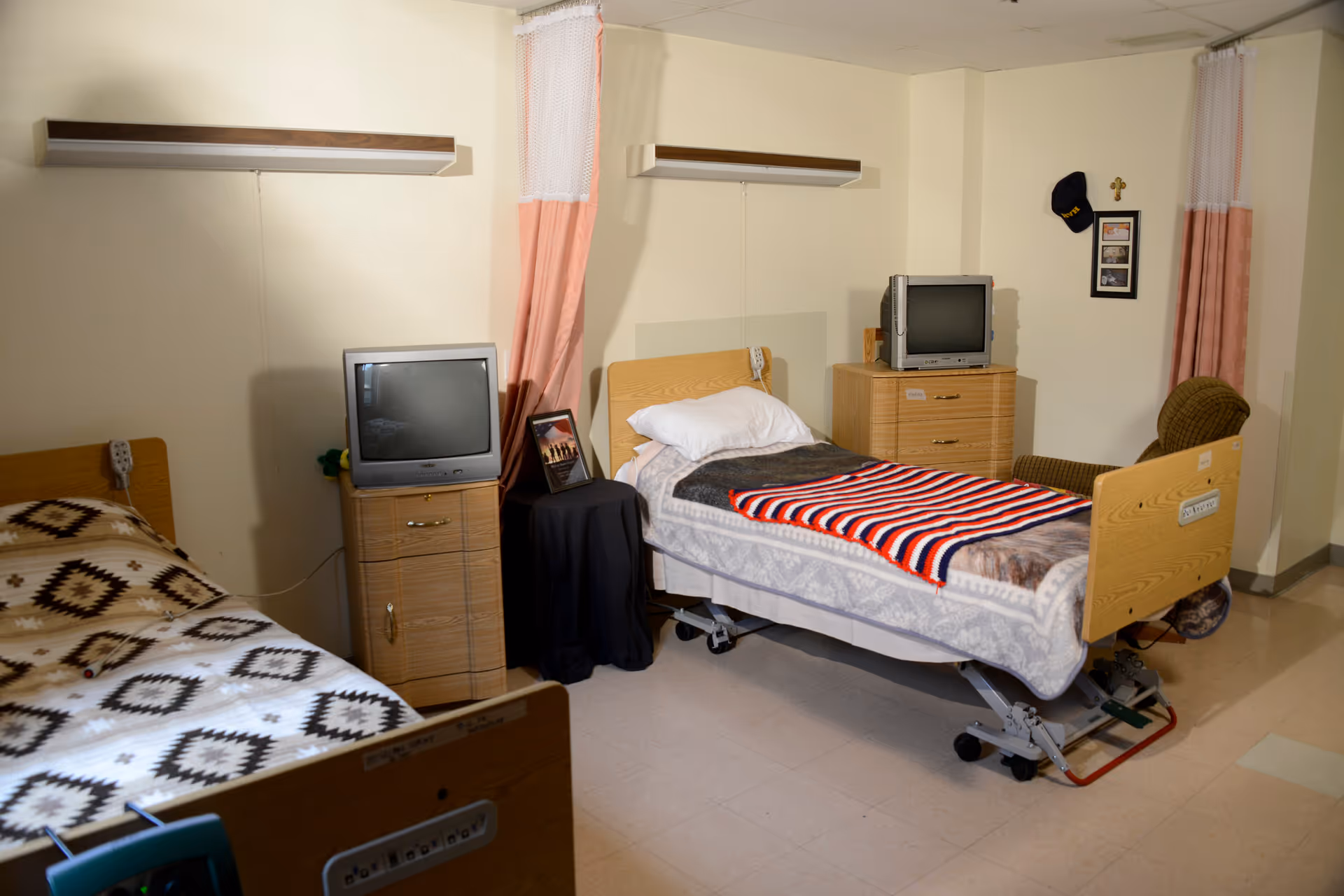 Shared patient bedroom with two adjustable hospital beds, bedside dressers, small TVs and privacy curtains.