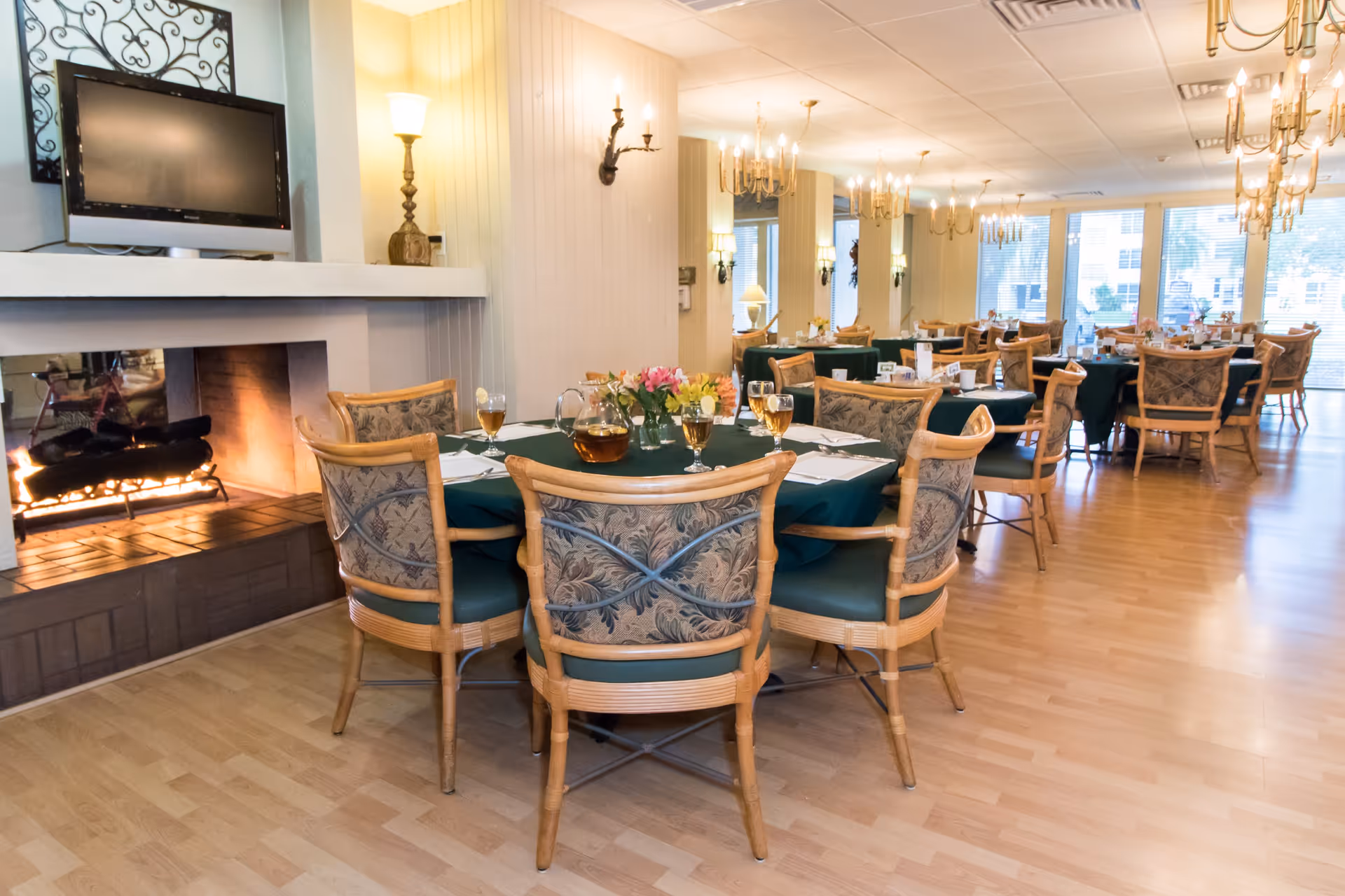 A dining room with multiple tables covered in dark green tablecloths, each set with glasses, napkins, and floral centerpieces. The room features wooden chairs with patterned cushions, a lit fireplace, a wall-mounted TV, and several chandeliers and wall sconces providing warm lighting. Large windows allow natural light to enter the space.