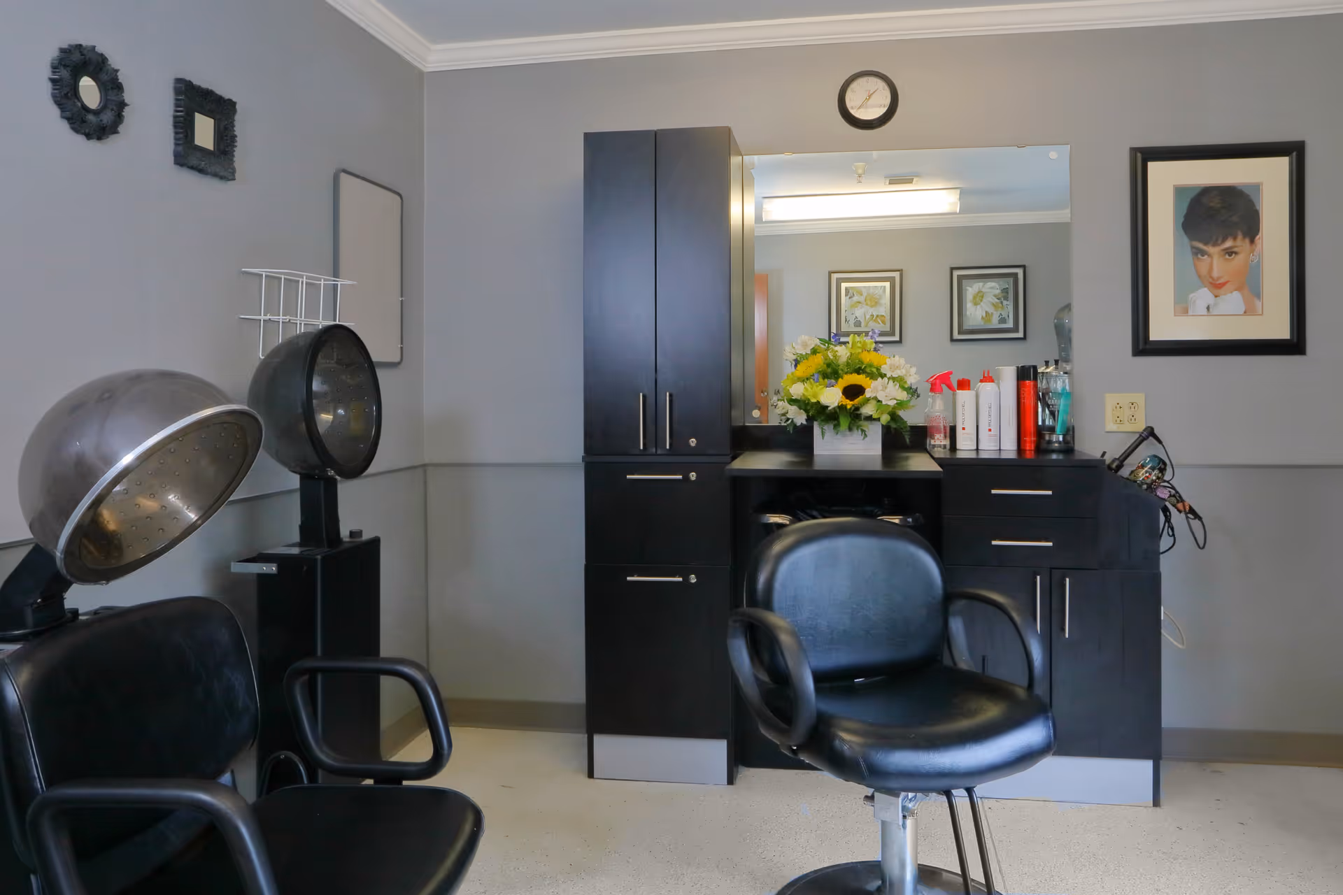 Interior of a hair salon area with two black salon chairs, two hair dryers, a black cabinet with a large mirror, various hair products on the counter, a clock above the mirror, and framed pictures on the walls.