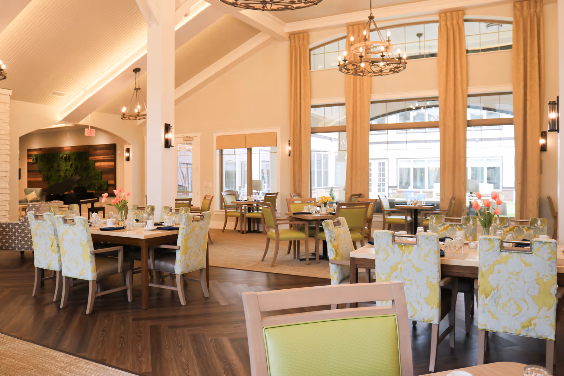 A bright and spacious dining room with several tables and chairs arranged neatly. The chairs have patterned upholstery in soft yellow and green tones. Large windows with beige curtains allow natural light to fill the room. There are chandeliers hanging from the high ceiling, and a grand piano is visible in the background near a wall with decorative wood and greenery.