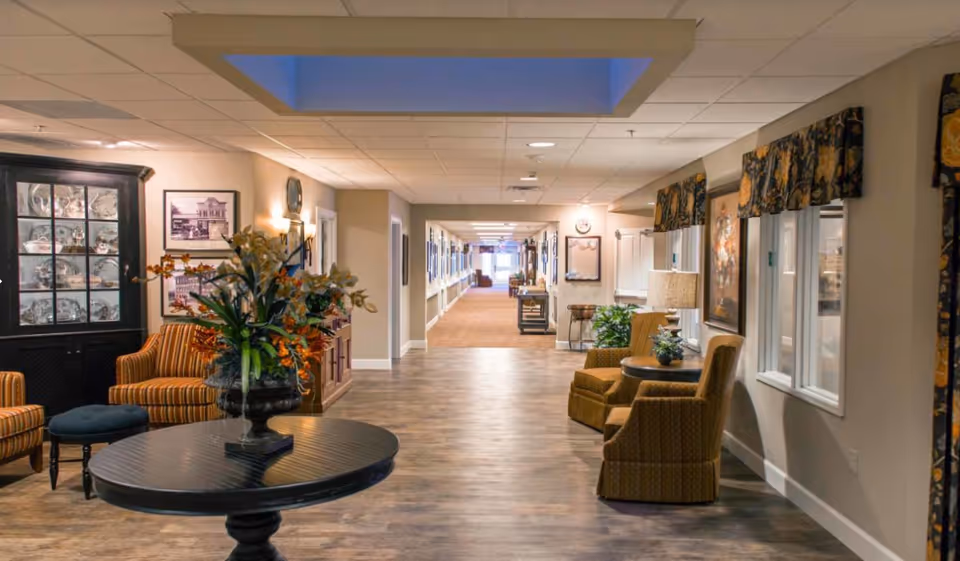 A well-lit interior hallway of a senior living facility with wooden flooring and beige walls. There are comfortable armchairs and small tables with lamps and plants along the walls. A round table with a floral arrangement is in the foreground. The hallway extends into the distance with framed pictures on the walls and a carpeted section further down.