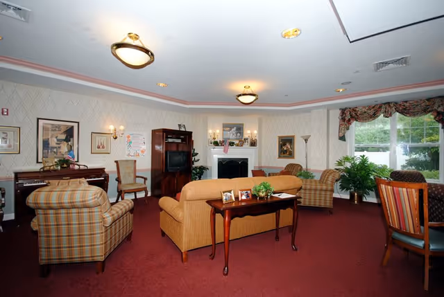 A cozy living room with patterned wallpaper, a red carpet, and various seating options including plaid armchairs and a beige sofa. There is a wooden table behind the sofa with framed photos and a small plant. The room features a fireplace with decorations on the mantel, a TV cabinet, framed artwork on the walls, and large windows with floral curtains letting in natural light. Several potted plants add greenery to the space.