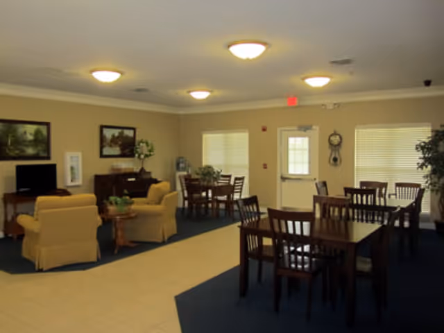 A well-lit common area in an assisted living facility featuring multiple wooden tables and chairs arranged for dining or socializing. There are two beige armchairs with a small round table between them, a television on a stand, framed artwork on the walls, and a clock near the exit door. The room has beige walls, blue carpet, and tile flooring near the seating area.