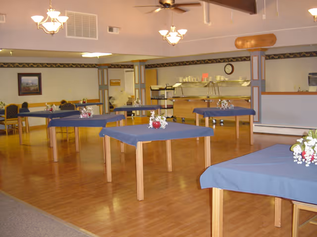 Spacious dining room with multiple square tables covered in blue tablecloths and small floral centerpieces and a buffet area in the background.