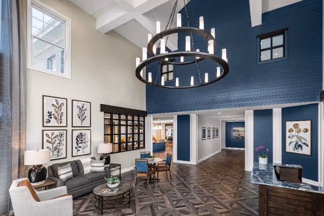 A spacious and well-lit common area with high ceilings featuring a large circular chandelier. The room has a dark blue accent wall with small windows near the ceiling, light beige walls, and patterned wooden flooring. There is a seating area with a gray sofa, two armchairs, a coffee table, and a small round table with four chairs. Decorative framed botanical prints hang on the wall, and a marble-topped reception desk with a potted plant is visible on the right side.