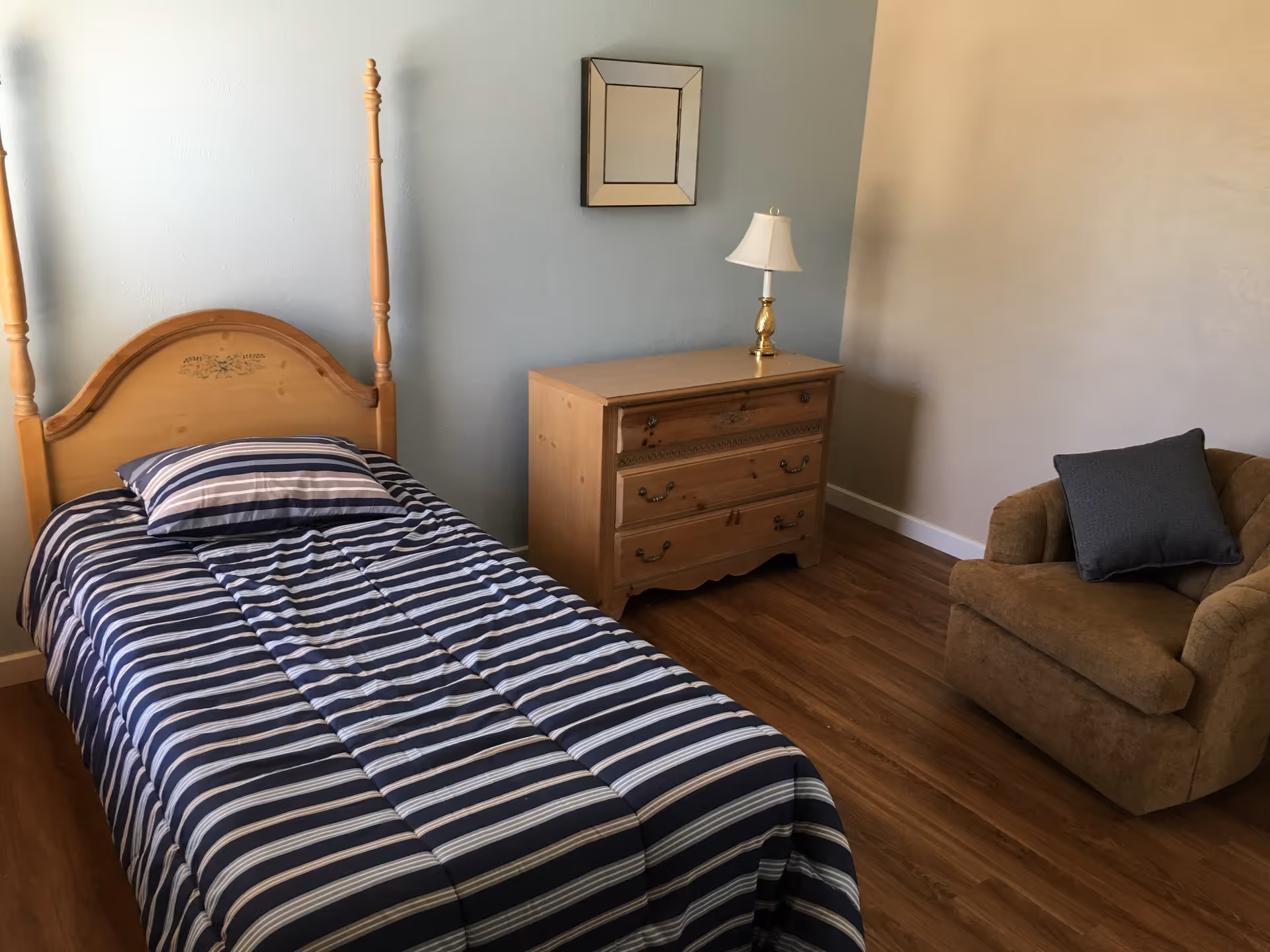 A bedroom with a wooden single bed featuring a striped blue and white comforter and matching pillow. Next to the bed is a wooden dresser with three drawers and a small lamp on top. A small square mirror hangs on the light blue wall above the dresser. To the right of the dresser is a brown upholstered armchair with a dark gray cushion. The floor is wooden.