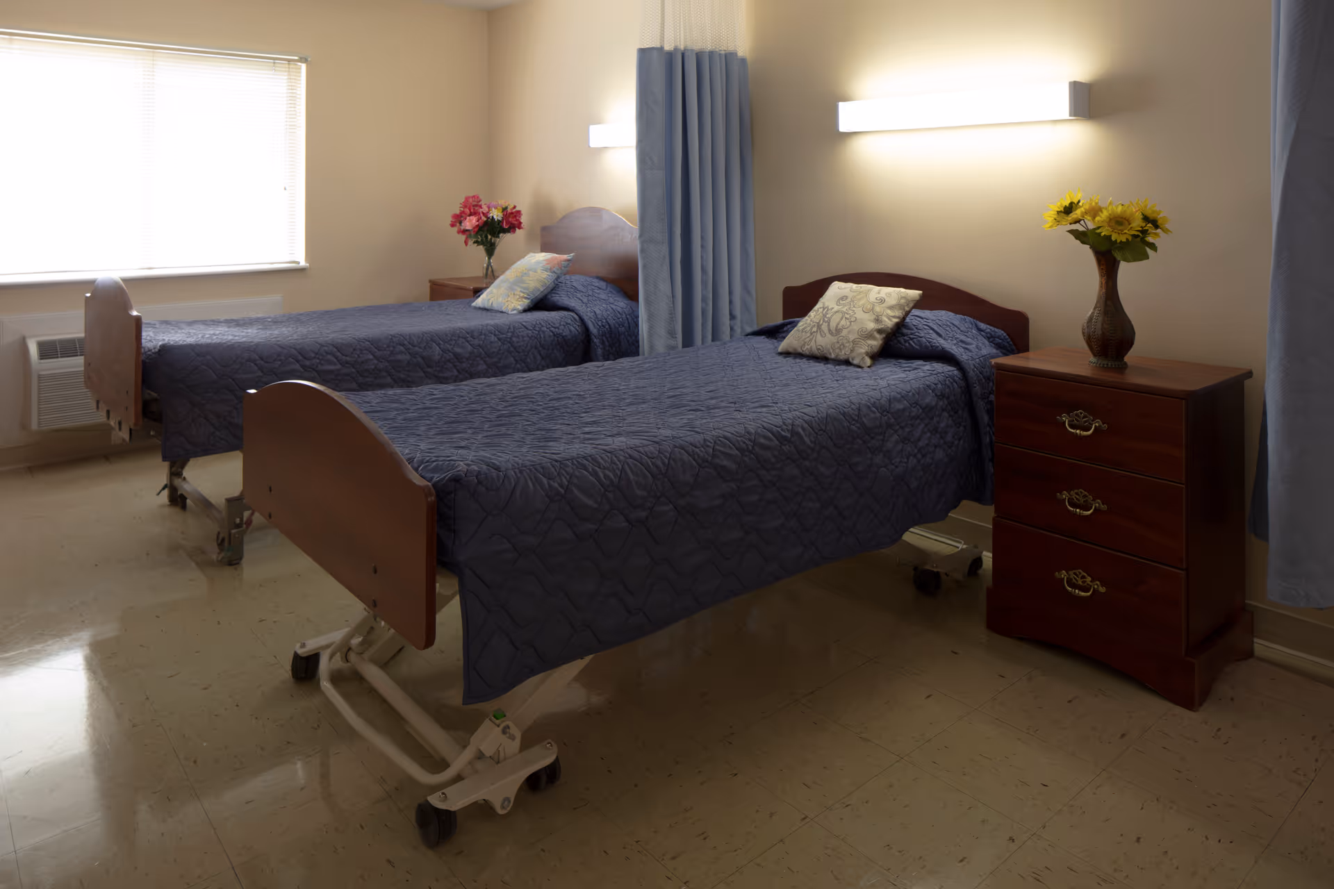 Two adjustable hospital-style beds with blue quilts in a shared senior facility bedroom with a nightstand and flowers.