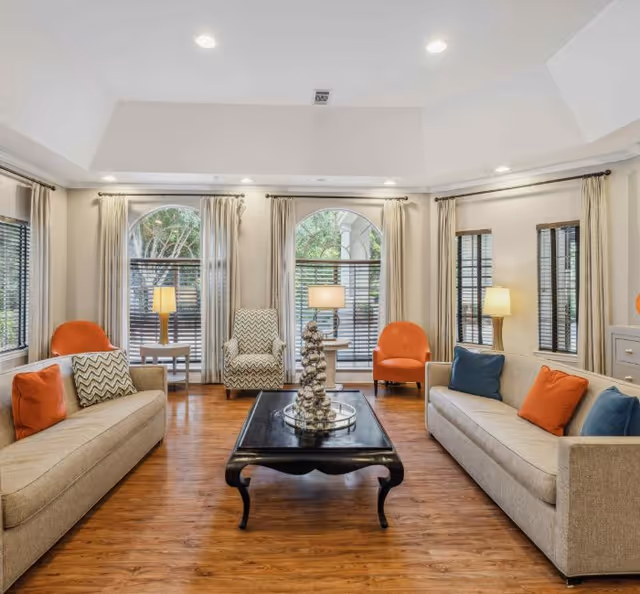 Bright senior living lounge with beige couches, colorful chairs, and arched windows.