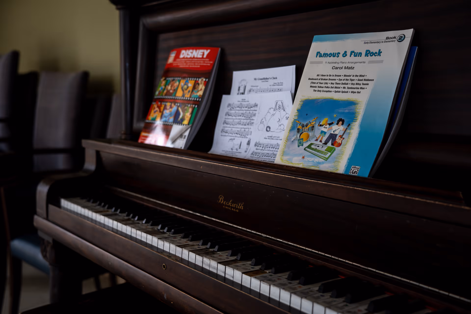 A close-up view of a dark wooden piano with sheet music and songbooks placed on the music stand. The visible songbooks include a Disney-themed book and a book titled 'Famous & Fun Rock'. In the background, there are a few chairs lined up against the wall.