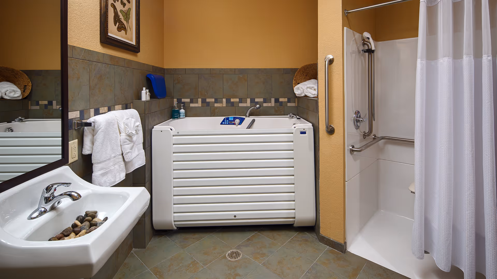 Accessible bathroom with a walk-in bathtub, a shower area with grab bars and a white curtain, a white sink with a faucet and decorative stones inside, a towel rack with white towels, and tiled walls and floor.