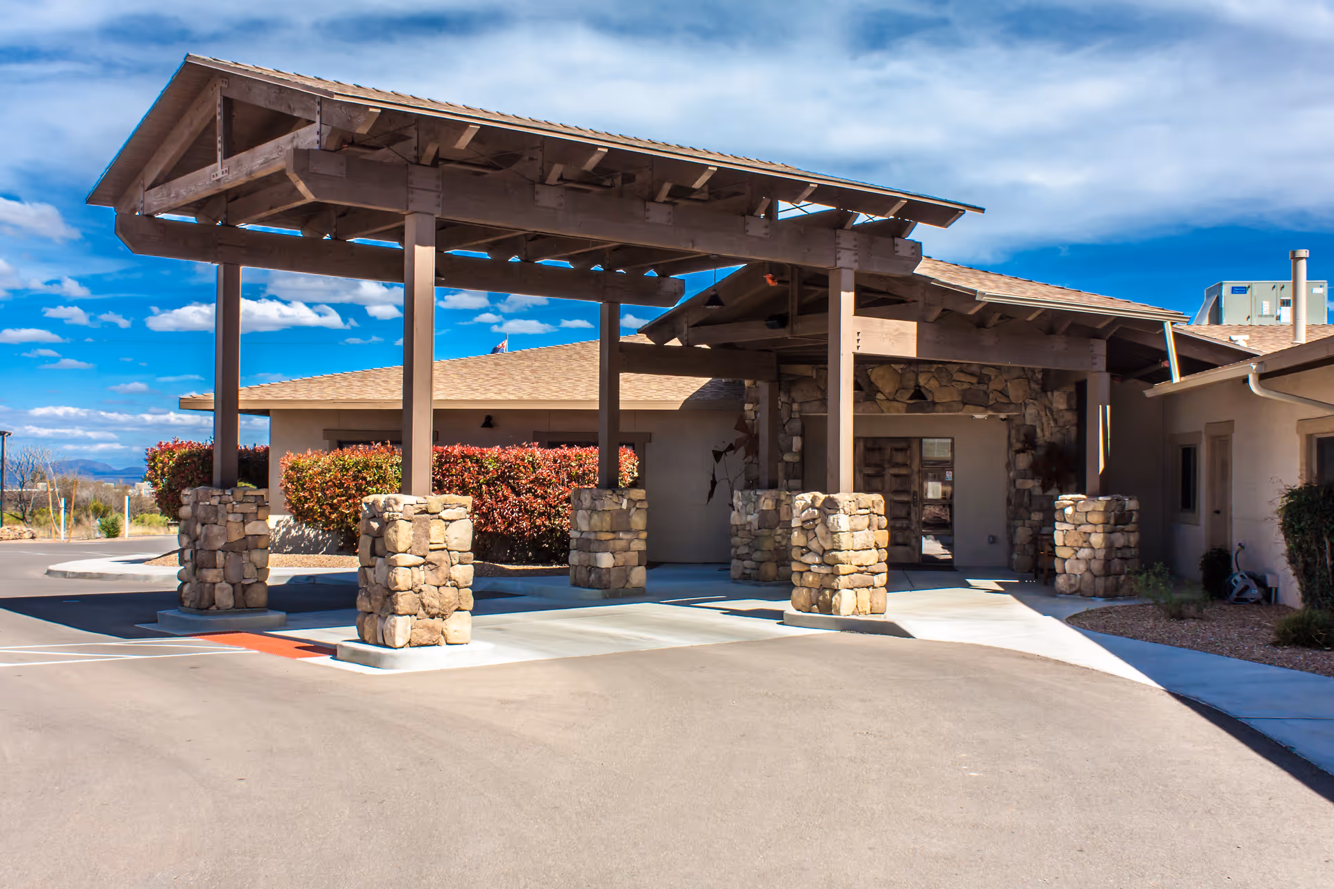 Entrance of Via Elegante Assisted Living Sierra Vista Canyons featuring a covered drop-off area supported by stone pillars, with a building and clear blue sky in the background.