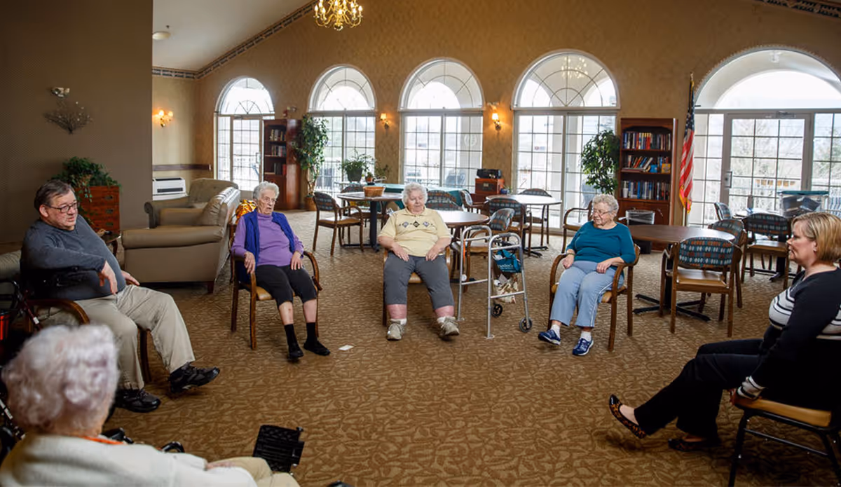 A group of elderly people sitting in a circle on chairs in a spacious, well-lit common room with large arched windows, carpeted floor, and several tables and bookshelves in the background. The room has warm lighting and an American flag is visible near the windows.