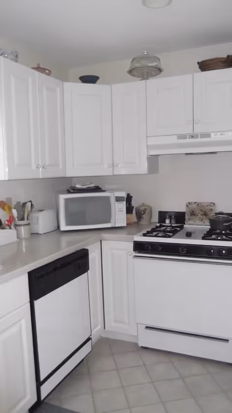 A compact kitchen with white cabinetry and appliances including a microwave, dishwasher, gas stove, and toaster. The countertops are light-colored, and there are various kitchen utensils and decorative items placed on the counters and above the cabinets.