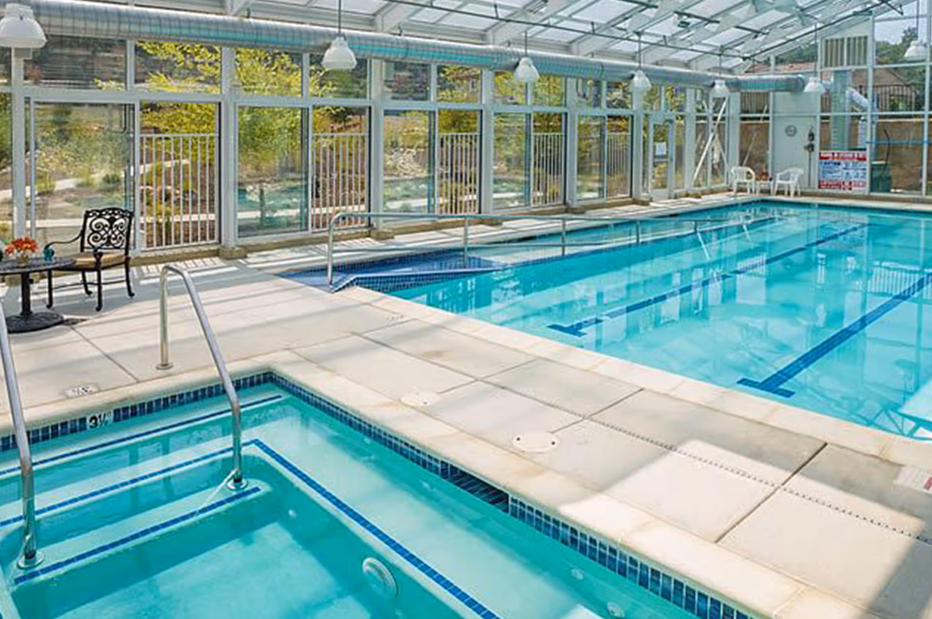 Glass-enclosed indoor swimming pool and adjacent hot tub with poolside seating and walkways.
