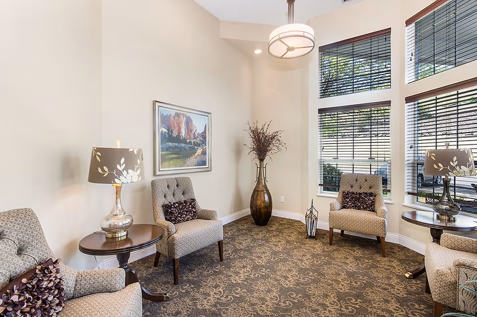 A cozy sitting area in an assisted living facility with four upholstered armchairs arranged around two wooden side tables, each with a decorative lamp. The room has large windows with blinds, a patterned carpet, a tall vase with branches in the corner, and a framed landscape painting on the wall.