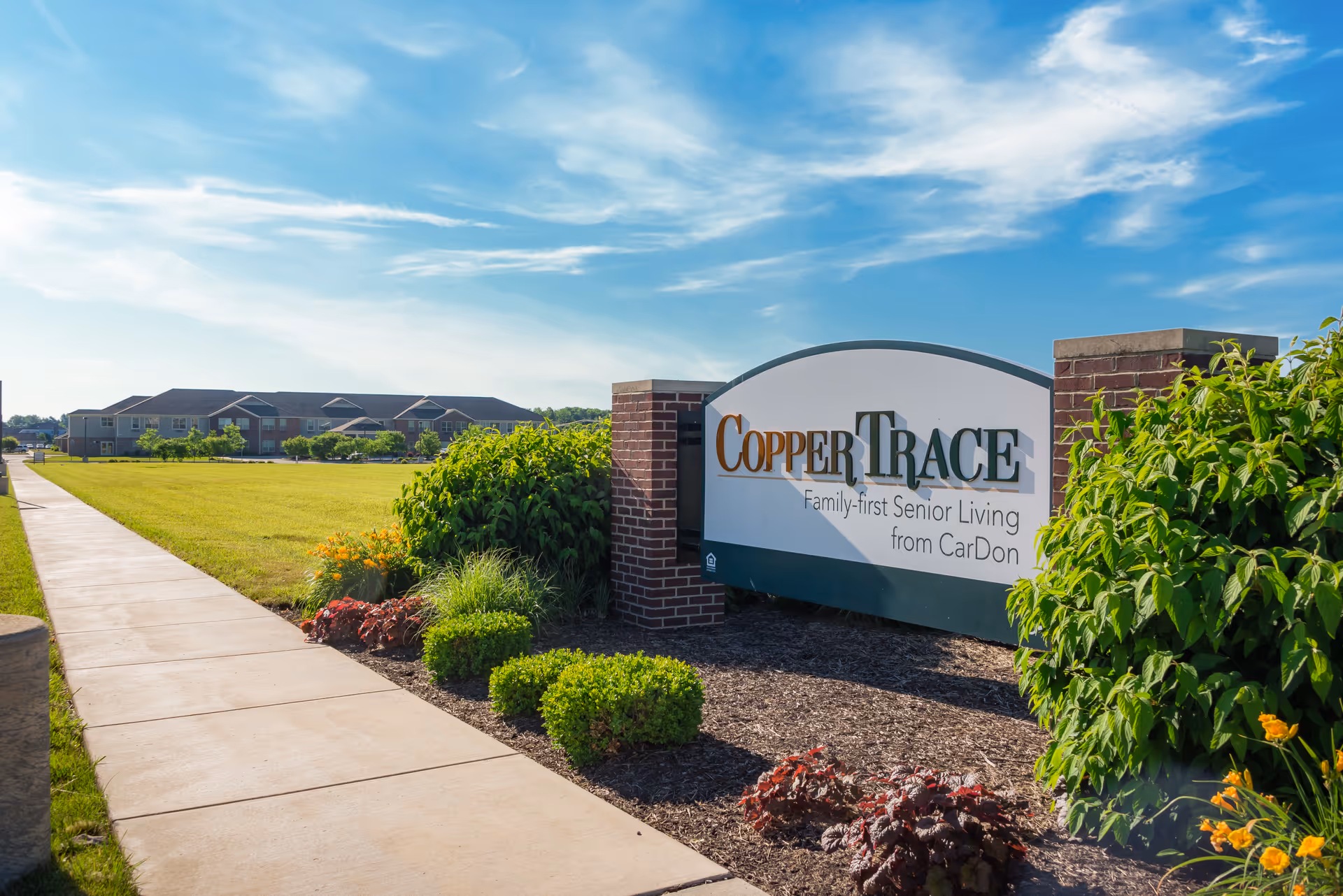 Entrance sign for Copper Trace Family-first Senior Living with landscaped grounds, a sidewalk, and the facility building under a blue sky.