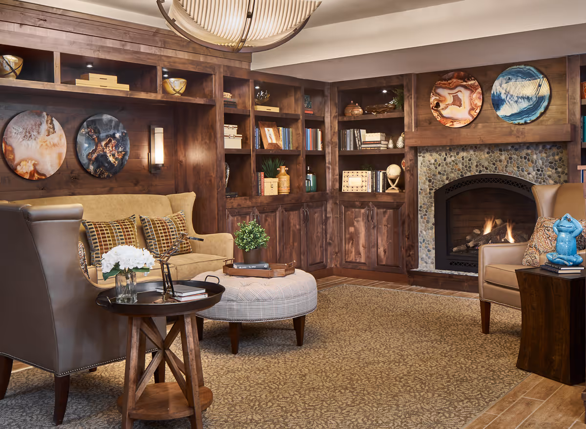 Cozy living room with a beige sofa, two armchairs, a round ottoman, and a small wooden side table with a vase of white flowers. The room features built-in wooden bookshelves filled with books and decorative items, a stone fireplace with a fire burning, and three round abstract art pieces mounted above the fireplace and sofa.