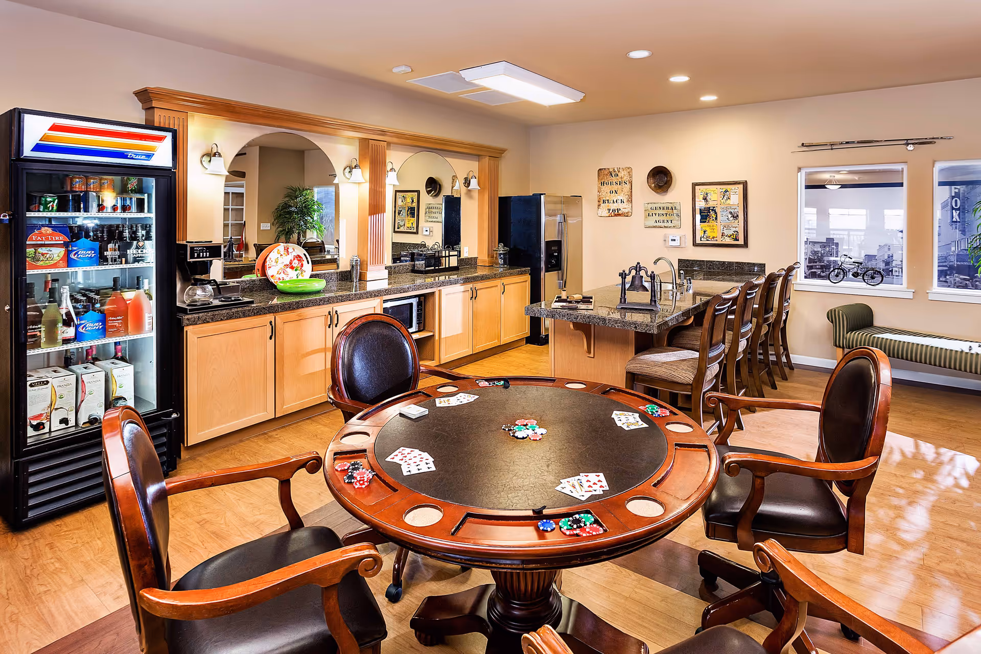A senior living facility common room featuring a round poker table with playing cards and poker chips, surrounded by four wooden chairs with leather seats. Behind the table is a kitchenette area with wooden cabinets, a granite countertop, a refrigerator, a microwave, and a beverage cooler stocked with drinks. There is a long counter with bar stools, wall decorations, and two windows with black and white cityscape images.