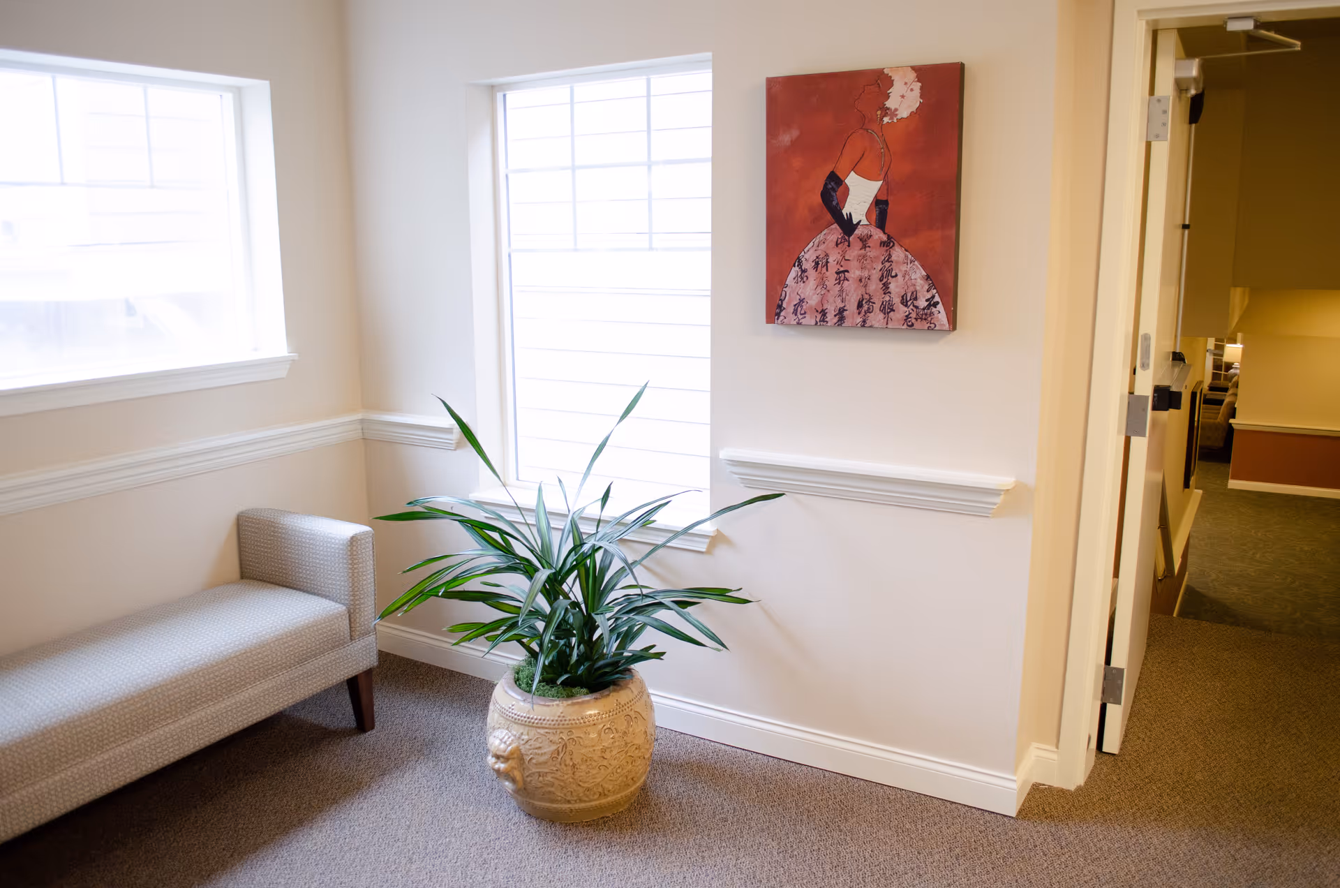 A bright corner of an interior space with two large windows, a patterned beige bench, a large decorative potted plant, and a painting of a woman in a red dress on the wall. The area has beige walls and carpeted floor, with a hallway visible through an open door.