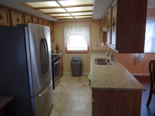 Galley kitchen with stainless steel refrigerator and stove, wooden cabinets, granite countertops with sink, and a window above a trash can.