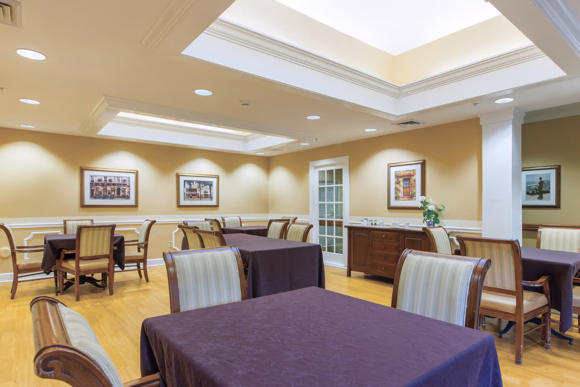 Dining room with several tables covered in dark tablecloths, wooden chairs with striped upholstery, framed artwork on beige walls, and skylights overhead.