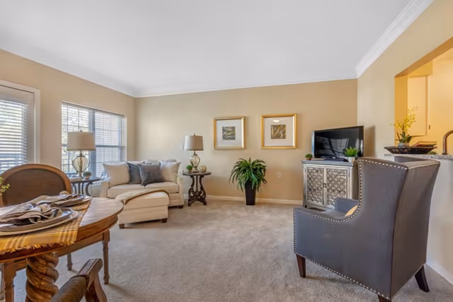 A bright living room with beige walls and carpet. The room features a cream-colored sectional sofa with gray and beige pillows, two side tables with lamps, a potted plant, a TV on a decorative cabinet, and a gray armchair with nailhead trim. There is a round wooden dining table with place settings in the foreground and two framed pictures on the wall.