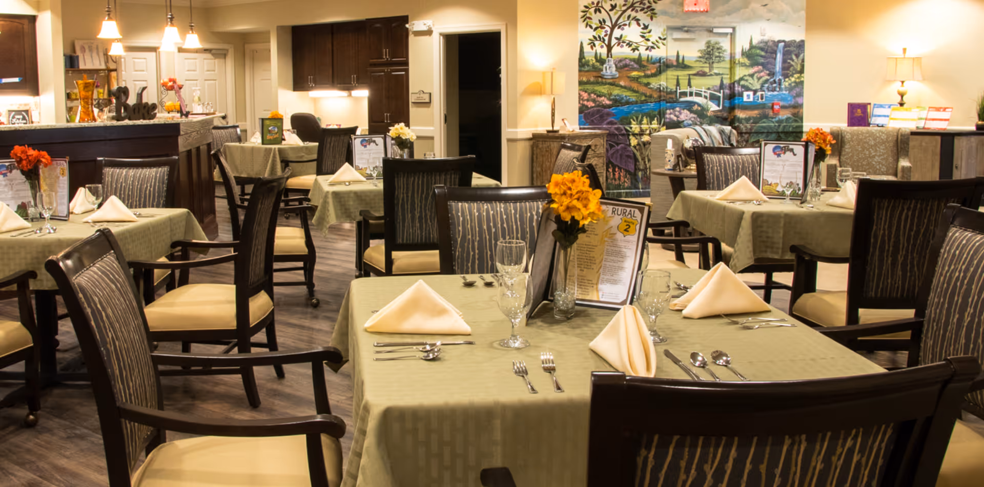 A dining room with several tables covered in green tablecloths, each set with folded napkins, glassware, silverware, and small flower arrangements. The room features wooden chairs with patterned upholstery, warm lighting, and a colorful mural on the back wall depicting a scenic landscape with trees, a bridge, and a waterfall.