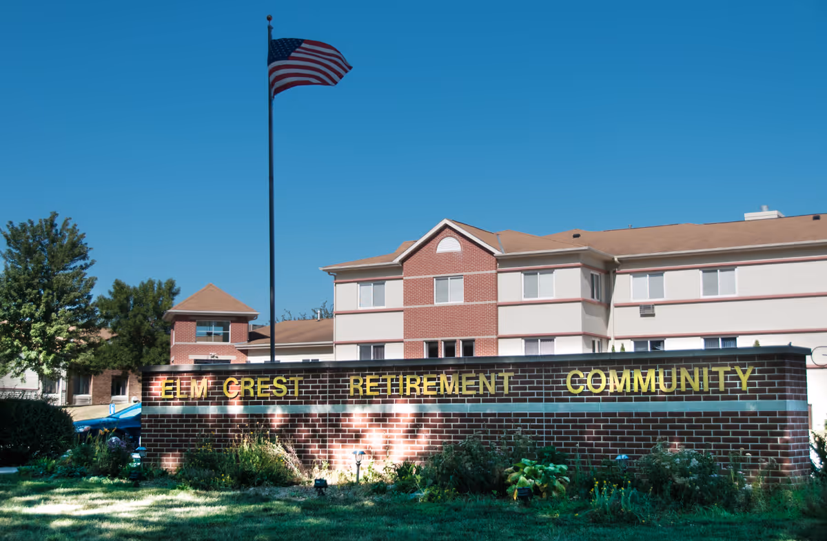 Exterior view of Elm Crest Retirement Community building with a brick sign in front displaying the community name. An American flag flies on a tall flagpole behind the sign, and there are trees and shrubs around the area under a clear blue sky.