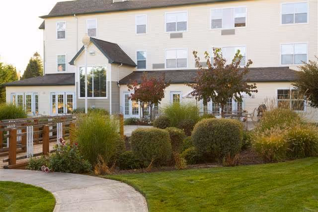 Exterior view of a multi-story senior living facility with beige siding, multiple windows, and a landscaped garden featuring trimmed bushes, small trees, and a curved concrete walkway leading to the entrance.