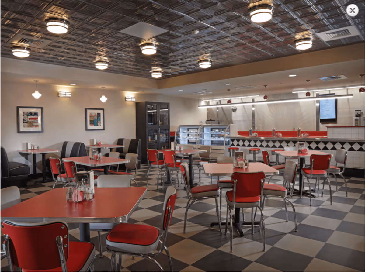 A retro-style dining room with red and gray chairs around tables with red tabletops. The floor has a black and white checkered pattern, and the ceiling features decorative metal tiles with round light fixtures. There is a counter area with a display case and a menu screen on the wall in the background.